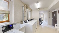 This shared amenity space is designed to serve the practical needs of its users, combining a fully equipped laundry room with a dedicated bicycle storage area. The laundry room is the centerpiece of the facility, featuring a row of full-sized, commercial-grade washing machines and dryers lining one wall, alongside a deep utility sink for hand-washing or soaking. The exposed white-painted brick walls lend a clean, industrial character to the space, while multiple windows with blinds allow natural light to brighten the interior. Overhead fluorescent lighting ensures clear visibility at all hours. A large folding table with seating provides a convenient workspace for sorting and folding laundry, and a wide mirror above the table adds a sense of openness. Nautical-themed framed artwork decorates the walls, adding a modest decorative touch. Adjacent to the laundry area, a dedicated bike storage room features sturdy wooden racks capable of accommodating multiple bicycles. This room is also enclosed by white-painted brick walls and benefits from natural light through its own windows. A separate utility area houses the building's mechanical and plumbing infrastructure. The light-colored flooring throughout ties the spaces together, creating a cohesive and functional facility.