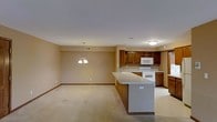 This single-floor space is designed with an open-concept layout that promotes a sense of spaciousness and easy flow between the kitchen and living areas. The kitchen is a standout feature, equipped with rich, warm-toned wooden cabinetry that provides ample storage. It includes a white electric range, a built-in microwave, and a double sink, all set against neutral countertops. A peninsula extends from the kitchen, offering additional workspace and serving as a natural divider between the kitchen and the adjoining living area. The living area is finished with light-colored carpet that adds warmth and comfort underfoot. Neutral, beige-toned walls run throughout the space, creating a cohesive and understated aesthetic that is easy to personalize. An elegant chandelier hangs above the living area, adding a decorative focal point and providing overhead lighting to complement the flush-mount ceiling fixtures found elsewhere in the space. Crown molding and baseboards frame the rooms with a clean, finished look. The overall ambiance is practical and welcoming, with a straightforward, well-organized layout that makes efficient use of the available space.