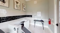 This single-level pet grooming space is thoughtfully designed to provide a clean, functional, and welcoming environment for pet care. The room features light grey walls that rise to notably generous ceiling heights, creating an open and airy atmosphere. Natural light floods the interior through multiple large windows, including a prominent multi-pane window that offers views of the surrounding greenery, as well as a glazed entry door that further brightens the space.

The walls are finished with a classic two-tone subway tile wainscoting — crisp white tiles topped by a bold band of glossy black tiles — adding a polished, professional aesthetic. Light-toned tile flooring runs throughout, contributing to the clean and easy-to-maintain character of the room.

Two built-in dog wash tubs are positioned along the tiled wall, each equipped with accessible ramps and hardware for efficient bathing. A professional grooming table with an adjustable arm is centrally positioned, providing a dedicated station for post-bath grooming. Whimsical framed dog portrait artwork adorns the walls, lending a lighthearted charm to the otherwise utilitarian space.

White baseboards and trim throughout complement the overall aesthetic, while HVAC venting ensures proper climate control. The combination of practical fixtures, durable finishes, and cheerful décor makes this an exceptional purpose-built grooming facility.