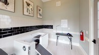 This single-level pet grooming space is thoughtfully designed to provide a clean, functional, and welcoming environment for pet care. The room features light grey walls that rise to notably generous ceiling heights, creating an open and airy atmosphere. Natural light floods the interior through multiple large windows, including a prominent multi-pane window that offers views of the surrounding greenery, as well as a glazed entry door that further brightens the space.

The walls are finished with a classic two-tone subway tile wainscoting — crisp white tiles topped by a bold band of glossy black tiles — adding a polished, professional aesthetic. Light-toned tile flooring runs throughout, contributing to the clean and easy-to-maintain character of the room.

Two built-in dog wash tubs are positioned along the tiled wall, each equipped with accessible ramps and hardware for efficient bathing. A professional grooming table with an adjustable arm is centrally positioned, providing a dedicated station for post-bath grooming. Whimsical framed dog portrait artwork adorns the walls, lending a lighthearted charm to the otherwise utilitarian space.

White baseboards and trim throughout complement the overall aesthetic, while HVAC venting ensures proper climate control. The combination of practical fixtures, durable finishes, and cheerful décor makes this an exceptional purpose-built grooming facility.