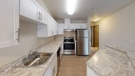 Located at 1007 115th Street Ct E in Tacoma, WA, this single-floor unit offers a well-organized layout connecting key living areas through a central hallway finished with light wood-look flooring. The kitchen is a functional highlight, featuring white shaker-style cabinetry extending to the ceiling, light stone-patterned countertops, a double-basin sink, and a full suite of stainless steel appliances including a refrigerator, range, and dishwasher. Ample counter space on both sides of the kitchen supports everyday meal preparation.

A dedicated laundry area near the entry provides in-unit washer accommodations alongside a tall storage cabinet with wood-grain paneling, adding practical convenience. The hallway connects to the bathroom, which includes white cabinetry, a vanity with stone-patterned counters, a lighted mirror, and a full tub with a stone-surround surround and curved shower rod.

The carpeted bedrooms offer a comfortable feel, with baseboard heating units for climate control. The larger bedroom benefits from two windows framing views of surrounding greenery, while a second bedroom provides a quieter, more enclosed atmosphere. The overall design maintains a clean, neutral palette throughout.