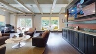 Located in Mechanicsville, Virginia, this single-story community space at Hanover Crossings offers a thoughtfully designed interior that balances architectural character with functional amenities. The main lounge area immediately impresses with its dramatic vaulted ceiling, accented by exposed decorative beams and a arched clerestory window that draws natural light deep into the space. Rich hardwood-style flooring runs throughout, providing a warm, cohesive foundation across all areas.

The open lounge features comfortable seating arrangements anchored by a sleek built-in fireplace, creating an inviting focal point beneath vibrant wall art. A dedicated workstation with desk and computer equipment sits nearby, offering a practical workspace alongside the relaxed seating. A secondary lounge area showcases coffered ceilings with painted beams, additional seating, a wall-mounted display screen, and a built-in navy cabinetry unit, with large windows framing lush outdoor greenery.

The kitchen area is well-appointed with dark cabinetry, a granite-topped island with bar seating, stainless steel appliances, a subway tile backsplash, and stylish pendant lighting. A casual dining nook with bar-height seating and ample window light completes the space, making it well-suited for both social gatherings and everyday use.