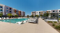This expansive outdoor amenity space is designed to deliver a resort-like experience within a contemporary multi-story residential community. At its centerpiece sits a generously sized swimming pool with clear blue water, surrounded by a wide, smooth concrete sun deck lined with rows of lounge chairs — both traditional recliners and sculptural wave-style loungers — offering ample space for relaxing under open skies.

Adjacent to the main pool is a dedicated spa, providing a more intimate retreat alongside the larger aquatic area. The deck transitions into a well-appointed outdoor lounge zone featuring modern pergola structures that provide shade over seating and dining arrangements, creating inviting spots for gathering throughout the day.

A built-in BBQ station with stainless steel grills is conveniently positioned near the pool perimeter, ideal for outdoor entertaining. The landscaping throughout the space incorporates drought-tolerant desert plantings, ornamental grasses, and decorative gravel beds, reflecting a low-maintenance yet visually appealing aesthetic well-suited to a warm, sunny climate.

The surrounding multi-story residential buildings, finished in a palette of warm wood tones, earthy grays, and muted purples, frame the courtyard on multiple sides, creating an enclosed, community-focused atmosphere. The entire space is secured with black metal fencing, ensuring privacy while maintaining an open, airy feel under clear blue skies.