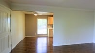 This single-floor space showcases a well-designed open layout that seamlessly connects the living area to the kitchen. The rich, dark hardwood floors run throughout, reflecting natural light and adding warmth to the interior. Crown molding lines the ceilings, lending a polished, classic finish to the space.

The living area is characterized by neutral, light-toned walls and clean white baseboards, creating a bright and airy atmosphere. A partial wall divides the living space from the adjoining area, providing subtle definition without closing off the open feel.

The kitchen is visible through the opening and features white cabinetry that contrasts attractively with the darker-toned accent wall behind it. A ceiling fan with a light fixture is mounted in the kitchen area, offering both ventilation and overhead lighting. A window in the kitchen fills the space with abundant natural light, offering a view of the greenery outside and enhancing the overall brightness of the interior.

The combination of classic architectural details, durable hardwood flooring, and an open, connected floor plan makes this a functional and inviting single-floor space.