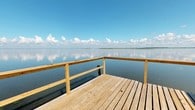 Located at 2200 East Bay Drive in Daphne, Alabama, this single-level waterfront property offers a rare combination of natural beauty and direct bay access. The standout feature of this property is its wooden dock, constructed with light-toned timber planking and sturdy railing that extends out over the calm, glassy waters of Mobile Bay. From the dock, residents and visitors are treated to expansive, unobstructed panoramic views stretching across the bay, where the sky and water meet in a seamless reflection of open blue horizons dotted with soft, billowing clouds. The setting conveys a profound sense of openness and serenity, characteristic of Alabama's Gulf Coast waterfront living. The dock provides direct water access, making it well-suited for boating, fishing, or simply enjoying the peaceful ambiance of bayside life. The natural light is abundant, with the wide-open water views ensuring sunlight throughout the day. The overall atmosphere of this property is calm and scenic, offering an idyllic coastal lifestyle defined by its immediate connection to the bay and the uninterrupted natural vistas that serve as a constant backdrop to everyday life.