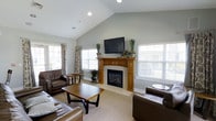 This single-story living space is defined by its vaulted ceilings, which lend an open, airy quality to the room. The soft gray-green walls provide a neutral and calming backdrop that complements the warm wood tones throughout the space. A central fireplace with a classic wood surround serves as the focal point of the room, topped by a wall-mounted flat-screen television and decorated with potted greenery, adding a natural touch to the setting. The fireplace surround features traditional craftsmanship with detailed column accents, contributing warmth and character to the overall design. Natural light flows generously into the space through multiple windows dressed with white plantation shutters and accented by patterned drapery panels, creating a bright yet comfortable atmosphere. The living area is furnished with rich, dark brown leather seating arranged around a wooden coffee table, offering a practical and cohesive layout. Neutral carpeting extends throughout the room, softening the space and enhancing its inviting feel. Recessed lighting in the ceiling complements the natural light, ensuring the room is well-lit at any time of day. Overall, the space balances classic design elements with a comfortable, functional arrangement.