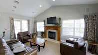 This single-story living space is defined by its vaulted ceilings, which lend an open, airy quality to the room. The soft gray-green walls provide a neutral and calming backdrop that complements the warm wood tones throughout the space. A central fireplace with a classic wood surround serves as the focal point of the room, topped by a wall-mounted flat-screen television and decorated with potted greenery, adding a natural touch to the setting. The fireplace surround features traditional craftsmanship with detailed column accents, contributing warmth and character to the overall design. Natural light flows generously into the space through multiple windows dressed with white plantation shutters and accented by patterned drapery panels, creating a bright yet comfortable atmosphere. The living area is furnished with rich, dark brown leather seating arranged around a wooden coffee table, offering a practical and cohesive layout. Neutral carpeting extends throughout the room, softening the space and enhancing its inviting feel. Recessed lighting in the ceiling complements the natural light, ensuring the room is well-lit at any time of day. Overall, the space balances classic design elements with a comfortable, functional arrangement.
