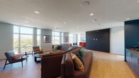 Located at 2439 Beatrice St in Dallas, TX, this contemporary single-floor communal space is thoughtfully designed to offer both comfort and visual interest. The main lounge area is anchored by a large, curved sectional sofa in warm brown tones, accented with colorful teal, rust, and patterned throw pillows that add a lively, curated character to the space. Recessed lighting throughout the smooth white ceiling provides consistent, well-distributed illumination across the open floor plan. Large, floor-to-near-ceiling windows line the walls, flooding the interior with abundant natural light and offering views of the surrounding Dallas skyline and cityscape. The wood-toned flooring adds warmth and continuity throughout. A secondary seating nook features a bold orange sectional sofa with teal accent pillows, framed artwork, and its own window view — offering an intimate retreat within the larger space. A striking feature wall finished in dark paneling anchors one end of the main lounge, creating a dramatic contrast against the lighter tones of the room. The kitchenette area is a standout, boasting floor-to-ceiling light blue tiles with copper-toned grout, deep teal lower cabinetry with brushed gold hardware, and a sleek stone countertop. The overall ambiance is modern, energetic, and welcoming.