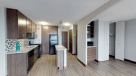 This single-floor unit offers a cohesive, modern design highlighted by light hardwood-style flooring that flows throughout the entire space, creating a unified and easy-to-navigate layout.

The kitchen serves as the functional heart of the unit, featuring rich dark-brown cabinetry that extends to ceiling height for generous storage. A mosaic tile backsplash adds visual texture behind the sink and gas range. Black appliances, including a refrigerator, gas range, built-in microwave, and dishwasher, provide a sleek, consistent finish. A central peninsula with a granite-style countertop offers an ideal space for casual dining or food preparation and helps define the kitchen from the adjacent areas.

A pass-through counter with a light granite surface further connects the kitchen to the living area, which is open and well-lit thanks to a pair of large windows dressed with vertical blinds. A baseboard heating system and wall-mounted HVAC unit ensure year-round comfort in the living space.

A built-in nook area provides additional storage and desk or display space, adding practical functionality near the main corridor.

The bathroom is clean and classically styled, featuring a pedestal sink, a framed medicine cabinet mirror, and wood-look flooring that complements the rest of the unit. Textured ceilings are present throughout, adding a consistent architectural detail across all rooms.