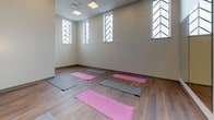 Located at 538-546 Broad Street in Newark, NJ, this dedicated yoga room offers a thoughtfully designed environment for focused movement and mindful practice. The space features warm-toned hardwood flooring throughout, providing a sturdy and inviting surface ideal for yoga and similar activities. A large wall of floor-to-ceiling mirrors spans one side of the room, allowing practitioners to monitor their form and creating a sense of expanded depth and openness. The room is framed by distinctive tall, narrow windows adorned with decorative chevron-patterned metalwork, which allow natural light to filter in while adding a unique architectural character to the space. Neutral, soft-toned walls contribute to a calming ambiance throughout. Recessed and panel lighting in the drop ceiling ensures consistent, even illumination. Wall-mounted sconce fixtures near the entry provide additional ambient lighting. The open floor plan accommodates multiple yoga mats laid out simultaneously, making it suitable for both group classes and solo sessions. An entry area with a secured door provides easy access and a clear transition into the practice space. Overall, the yoga room strikes a balance between functional design and a serene atmosphere, offering an inviting setting for wellness activities in the heart of Newark.