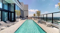 This elevated amenity floor offers a compelling combination of outdoor and indoor spaces designed for both relaxation and productivity, all set against a dramatic urban backdrop. The centerpiece of the outdoor area is a generously proportioned lap pool, its clear turquoise water flanked by clean-lined pool decking, white lounge chairs, and sculptural planters housing mature trees that add a natural contrast to the modern architectural aesthetic. The surrounding terrace is enclosed by sleek metal railings, opening up unobstructed panoramic views of a major city skyline — identifiable by prominent high-rise towers stretching to the horizon in every direction. Multiple seating vignettes are thoughtfully arranged throughout the terrace, offering intimate settings for outdoor gatherings with city views as the backdrop. Floor-to-ceiling glass curtain walls frame the transition from the outdoor terrace to the interior, flooding the inside with abundant natural light. The interior includes a sophisticated conference room featuring soaring ceilings, plush carpeting, a solid wood meeting table surrounded by contemporary black chairs, and a wall-mounted display screen. The overall design is cohesive and modern, balancing open-air leisure with functional indoor amenities at an impressive elevation above the city.