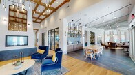 Located at 2568 E Glenn Ave in Auburn, Alabama, this single-floor community clubhouse delivers an impressive blend of modern design and welcoming ambiance. The space is defined by its soaring ceilings with exposed wood coffered beams and an abundance of natural light streaming through oversized windows and floor-to-ceiling glass sliding doors.

The main lounge area features rich hardwood flooring, plush navy blue velvet accent chairs, and a built-in entertainment console with cabinetry, creating an inviting social atmosphere. A striking wet bar area showcases bold blue cabinetry with brass hardware set against a herringbone-patterned tile backsplash, complemented by open wood shelving and a large mounted display screen.

Adjacent gathering spaces include a dining area with a long communal table, open metal shelving with decorative tile accents, and pendant lighting throughout. A casual seating nook with marble-top tables and large windows provides serene views of the outdoor pool area.

The lounge extends seamlessly to the outdoors through expansive glass doors, revealing a resort-style pool and patio. Throughout the space, layered lighting—including cascading pendant chandeliers and wall sconces—enhances the refined yet comfortable character of this thoughtfully appointed clubhouse.
