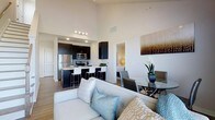 This contemporary two-story residence showcases a thoughtfully designed open-concept layout that effortlessly connects the main living areas. Soaring ceilings in the main living space create an impressive sense of openness and volume, filling the room with an airy, expansive ambiance.

The kitchen is outfitted with dark cabinetry that provides a striking contrast against light countertops, and features stainless steel appliances including a refrigerator, microwave, and range. A breakfast bar with seating extends from the kitchen island, making it ideal for casual dining or entertaining.

Adjacent to the kitchen, the dining area accommodates a stylish glass-top table with modern upholstered chairs, offering a dedicated space for formal meals. The living room is anchored by a comfortable neutral-toned sofa accented with decorative pillows, and the warm-toned hardwood-style flooring flows seamlessly throughout the main level, tying all areas together cohesively.

The overall design aesthetic is clean and modern, with a neutral color palette of whites and warm tones complemented by bold dark cabinetry accents. The open sightlines between the kitchen, dining, and living areas make the space feel connected and highly functional, ideal for both everyday living and entertaining across its two floors.