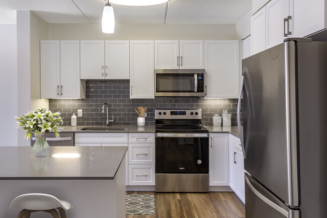 Renovated Package II kitchen with grey quartz countertops, white shaker cabinetry, stainless steel appliances, grey tile backsplash, and hard surface flooring throughout - Avalon Natick