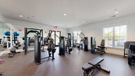 This fitness center is thoughtfully designed to support a full range of workout routines within a bright, well-organized single-level space. The interior features white walls accented by crown molding, recessed lighting, and speckled rubber flooring that provides both durability and comfort underfoot. Large windows fitted with plantation-style blinds line the walls, allowing natural light to filter throughout the space while offering views of the surrounding outdoor greenery. Glass-paneled entry doors further enhance the sense of openness.

The strength training area is well-appointed with multiple selectorized cable and resistance machines suited for targeted muscle group training, including chest press, lat pulldown, leg press, and seated row equipment. A dedicated free weights section houses a full rack of dumbbells in graduated weights, while a separate rack displays an assortment of kettlebells and medicine balls in various sizes for functional training. A Smith machine with an accompanying flat bench provides additional barbell training options.

Floor-to-ceiling mirrors are positioned strategically throughout the space, visually expanding the room and allowing users to monitor their form during workouts. The overall atmosphere is clean, organized, and functional, making it a practical and inviting environment for fitness enthusiasts of all levels.
