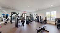 This fitness center is thoughtfully designed to support a full range of workout routines within a bright, well-organized single-level space. The interior features white walls accented by crown molding, recessed lighting, and speckled rubber flooring that provides both durability and comfort underfoot. Large windows fitted with plantation-style blinds line the walls, allowing natural light to filter throughout the space while offering views of the surrounding outdoor greenery. Glass-paneled entry doors further enhance the sense of openness.

The strength training area is well-appointed with multiple selectorized cable and resistance machines suited for targeted muscle group training, including chest press, lat pulldown, leg press, and seated row equipment. A dedicated free weights section houses a full rack of dumbbells in graduated weights, while a separate rack displays an assortment of kettlebells and medicine balls in various sizes for functional training. A Smith machine with an accompanying flat bench provides additional barbell training options.

Floor-to-ceiling mirrors are positioned strategically throughout the space, visually expanding the room and allowing users to monitor their form during workouts. The overall atmosphere is clean, organized, and functional, making it a practical and inviting environment for fitness enthusiasts of all levels.