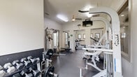 Located at 3501 E Gore Blvd in Lawton, Oklahoma, this fitness center is a thoughtfully designed facility spread across a single floor. The space is divided into two dedicated exercise rooms, offering a comprehensive workout environment for users of all fitness levels.

The primary exercise room is generously proportioned, featuring durable dark rubberized flooring that provides both safety and support during workouts. Cardio equipment, including multiple treadmills and a stair climber, lines one side of the room, positioned beneath windows that look out toward an outdoor pool area, allowing natural light to filter in. Ceiling fans maintain airflow and comfort throughout. A wall-mounted television adds entertainment for those engaged in longer cardio sessions.

The strength training area is equally well-appointed, housing a cable machine system, a weight bench, a dumbbell rack, and various resistance machines including a multi-function cable trainer. A two-tone wall treatment with a dark lower half gives this zone a focused, gym-like atmosphere.

A secondary, quieter exercise room offers a more intimate setting, featuring a recumbent bike, a foldable workout board, and a row of windows that bathe the space in soft, natural light. Clean white walls and dark flooring keep the aesthetic consistent and functional throughout the entire facility.