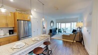 Located at The Shipyard At Port Jeff Harbor in Port Jefferson, NY, this thoughtfully designed residence combines modern style with comfortable living. The open-concept floor plan seamlessly connects the kitchen, dining, and living areas, creating a bright and inviting atmosphere throughout. The kitchen features stainless steel appliances, warm wood cabinetry, and a granite-topped island with bar seating — perfect for casual dining or entertaining. Stylish pendant lighting overhead adds a contemporary touch. Rich hardwood flooring flows throughout the main living area, grounding the space with warmth and continuity. The living room is generously proportioned, bathed in natural light streaming through large sliding glass doors that open to a private balcony with views of the surrounding harbor area. The bedroom, visible through a connecting doorway, offers a cozy and private retreat. The overall ambiance is modern yet welcoming, with clean white walls, thoughtful décor accents, and an airy feel that reflects the coastal character of Port Jefferson Harbor. This residence offers an exceptional combination of interior comfort and scenic waterfront surroundings.