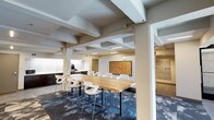 This single-floor communal space combines modern design with functional amenities, creating a versatile environment suited for both social interaction and focused work. The open layout is anchored by a long butcher-block communal table surrounded by contemporary white bar-height chairs, positioned beneath an exposed beam ceiling with linear LED lighting that adds an industrial character to the space.

The flooring features a striking patterned carpet with blue, grey, and white tones, interspersed with large-format tile sections that help define distinct zones throughout the space. A well-appointed kitchenette lines one wall, outfitted with dark cabinetry, white countertops, a commercial-grade coffee machine, water dispenser, beverage refrigerator, and a sink — providing comprehensive refreshment options.

Multiple seating areas create varied environments within the space. A lounge nook near a window features warm-toned accent chairs and natural light, while a separate area near the entrance offers a pair of blue upholstered chairs alongside a floor lamp and framed wall art, contributing a welcoming residential feel. A bank of mailboxes and dual access doors are also visible, suggesting a residential building amenity or lobby-level common area.