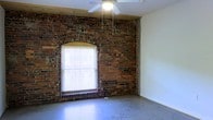 This single-floor space showcases a compelling blend of historic architectural character and raw industrial aesthetic. The most prominent feature is the exposed brick wall, which spans a significant portion of the interior and displays rich, aged texture with varied tones of red and brown. Set within this brick wall is a classically styled arched window opening, framing a standard double-hung window fitted with horizontal blinds that allow for natural light control. The window provides an infusion of natural light, illuminating the open interior. The flooring consists of bare concrete, reinforcing the industrial character of the space. Overhead, a ceiling fan is centrally mounted, offering both air circulation and a functional focal point. A wooden beam detail runs along the upper edge of the brick wall, providing a natural transition between the historic masonry and the white drywall that lines the remaining walls. The contrast between the textured brick and the smooth white walls creates a dynamic visual balance, giving the space a versatile character well-suited to a variety of uses. The overall atmosphere is open, raw, and full of architectural potential.