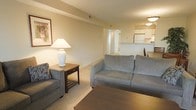 This single-floor residence offers a thoughtfully arranged open-concept layout that connects the living area to the kitchen, creating a cohesive and functional living environment. The living room is furnished with plush grey upholstered seating, including a sofa and armchair, complemented by a wooden side table topped with a classic white ceramic lamp. A framed artwork on the wall and a patterned accent pillow add subtle decorative character to the space. Soft, neutral-toned walls and wall-to-wall carpeting contribute to a warm and understated ambiance throughout.

A low breakfast bar or counter visually separates the living area from the kitchen beyond, which is outfitted with white cabinetry and appliances, including a refrigerator. Overhead lighting in the kitchen area provides a bright contrast to the more softly lit living space. A hallway visible in the background suggests additional rooms beyond, offering a sense of depth and privacy within the layout. The overall design is practical and comfortable, with a clean, neutral palette that creates a welcoming and livable atmosphere.