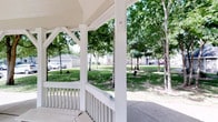 This gazebo is a classic open-air structure finished in crisp white, featuring sturdy decorative columns and a traditional pitched roof supported by angled brackets that add architectural character. The perimeter is lined with a white balustrade railing, providing a defined yet open boundary that maintains clear sightlines to the surrounding landscape.

The structure opens directly to a well-maintained green space filled with mature trees, whose canopies provide natural shade and a sense of enclosure. The lush lawn extends outward in multiple directions, creating a park-like setting that enhances the gazebo's role as a peaceful outdoor gathering spot. A concrete pathway connects the gazebo to the broader grounds, offering easy accessibility.

Natural light filters through the tree cover, creating a dappled, comfortable atmosphere throughout the day. The surrounding environment includes nearby buildings visible through the treeline, suggesting the gazebo serves as a communal amenity within a larger residential or mixed-use setting. Overall, this space offers a tranquil, shaded outdoor retreat well-suited for relaxation or informal gatherings.