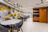 Located at 50 Portland Street in Toronto's vibrant King West neighbourhood, this contemporary amenity lounge offers a well-appointed, single-level space designed for both relaxation and productivity. The room immediately draws the eye with a striking full-height feature wall adorned in a bold geometric pattern of yellow, grey, and white triangles, adding a dynamic visual anchor to the otherwise clean, modern interior.

Large-format white marble-look porcelain floor tiles extend throughout, contributing to a bright and polished aesthetic. Recessed linear LED lighting runs across the white ceiling, complemented by a sculptural multi-arm pendant fixture that adds an artistic touch to the seating area.

Multiple round marble-top tables paired with contemporary grey shell chairs provide comfortable, flexible seating for casual gatherings or individual use. Along one wall, a built-in wooden counter with mounted flat-screen monitors and individual workstations offers a dedicated area for focused work.

A warm-toned wooden built-in bookshelf stocked with books and a printer provides a resourceful library nook. The kitchenette area features a concrete-finish counter, undermount sink, under-counter refrigerator, microwave, and coffee station, making the space fully self-sufficient. Sheer and fabric drapery softens the perimeter, completing this thoughtfully designed communal lounge.