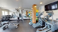 This fitness center is thoughtfully organized into distinct zones, offering a comprehensive range of equipment and amenities for varied workout routines. The main strength and cardio area features durable rubber flooring and recessed lighting, housing a multi-function cable machine, adjustable weight bench, elliptical trainers, and a full rack of medicine balls and stability equipment. Wall-to-wall mirrors amplify the sense of space while allowing users to monitor their form. A mounted flat-screen television provides entertainment during workouts.

A dedicated cardio room accommodates multiple treadmills side by side, each positioned beneath wall-mounted televisions, with a window offering outdoor views. An adjacent area provides additional strength-training machines, including leg press and rowing equipment, arranged beneath windows that allow natural light to fill the space.

The facility also includes a separate studio room designed for yoga, stretching, and group classes. This bright, open area features light hardwood flooring, a drop ceiling with panel lighting, floor-to-ceiling mirrors along one wall, ballet-style wall-mounted bars, and windows with exterior views. Yoga mats are available for use, making this a versatile space for low-impact and flexibility-focused training.