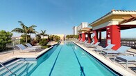 This exceptional outdoor amenity space offers a resort-like experience defined by a long, pristine lap pool with clearly marked lanes and shimmering turquoise water. The pool is flanked on both sides by a generously sized sun deck, lined with sleek lounge chairs ideal for relaxing in the Florida sunshine. Lush palm trees are thoughtfully integrated throughout the surrounding landscape, adding a tropical aesthetic that complements the warm, coastal atmosphere. A covered pavilion anchors one side of the deck, featuring a bold red and white color scheme with a clay tile roof, providing shaded seating and housing what appears to be a pool service or safety station. The open-sky setting allows for abundant natural light throughout the day, while sweeping views of the surrounding urban skyline — including high-rise towers — lend the space a dynamic, cosmopolitan energy. The clean, well-maintained surfaces and structured layout reflect a facility designed for both active use and leisurely enjoyment. This space is well-suited for lap swimming, sunbathing, and social gatherings, making it a standout amenity within its larger complex.