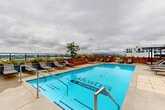 Located atop a modern residential building in LaSalle, Québec, these rooftop common areas offer a well-appointed retreat for residents. The interior includes a stylish combined kitchen and living room, where a large island with a dark countertop and integrated cooktop anchors the space beneath a stainless steel range hood. Dark open shelving, full-size appliances, and a warm wood dining table with seating complete the kitchen zone. The adjacent living room centers on a striking dark wood feature wall with a linear fireplace flanked by built-in shelving, with direct access to the outdoor terrace through glass French doors. The dining room provides a versatile gathering area, illuminated by natural light through large windows and doors, with views onto the rooftop terrace. Outside, the expansive terrace is equipped with multiple BBQ stations beneath a pergola with string lighting, ample seating at cafe-style tables, and a stunning rooftop swimming pool surrounded by lounge chairs. Glass railings along the perimeter frame sweeping panoramic views of the LaSalle skyline and surrounding urban landscape, making this rooftop an exceptional shared amenity space.