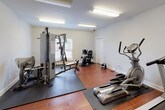 Located at 100 Estella Ct in Florence, Alabama, this single-floor fitness center is a well-equipped exercise facility designed to support a comprehensive workout routine. The space features warm hardwood-style flooring throughout, crisp white walls, and bright overhead fluorescent lighting that ensures excellent visibility across the entire room.

The cardio area is equipped with multiple treadmills and elliptical trainers, each placed on protective rubber mats, along with a stair stepper and a wall-mounted television for entertainment during workouts. On the strength training side, a large multi-function cable and weight machine takes center stage, accompanied by a recumbent stationary bike and additional resistance equipment, providing options for a full-body strength regimen.

Natural light filters in through windows fitted with white horizontal blinds, adding a bright and open feel to the space. Protective rubber floor mats are strategically placed beneath the heavier equipment to preserve the flooring and provide stability. A yoga mat is also available for stretching and floor exercises. The room includes a dedicated door for entry and exit, and a paper towel dispenser is mounted on the wall for convenience. The overall layout is organized and spacious, allowing multiple users to exercise comfortably at the same time.