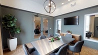 This single-floor space presents a thoughtfully designed meeting or conference room with a refined, contemporary aesthetic. The room is anchored by a sleek metallic rectangular table surrounded by upholstered herringbone-patterned chairs, offering comfortable seating for a group of six or more. Walls finished in a sophisticated slate blue-gray tone lend a calm, professional ambiance, complemented by crisp white crown molding and ceiling details that add architectural depth.

Overhead, a striking globe-style chandelier with exposed Edison bulbs serves as a focal point, blending industrial and modern design sensibilities. Recessed lighting supplements the chandelier, ensuring the space is well-lit throughout. A wall-mounted flat-screen television is positioned for easy viewing during presentations or collaborative sessions, accompanied by a warm wood credenza beneath it.

A window brings in natural light, enhancing the inviting atmosphere of the room. Glass-paneled French doors connect the space to an adjacent lounge or common area, which features exposed brick accents and warm lighting, suggesting a versatile, multi-use facility. Light wood-toned flooring with a parquet-style pattern runs throughout, tying together the room's cohesive and polished interior design.