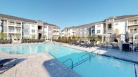 This single-story residence is part of a well-appointed multi-building community designed with a cohesive exterior aesthetic, featuring gray and white facades with balconies and manicured landscaping. A standout amenity of the community is the expansive resort-style swimming pool, surrounded by a tiled deck lined with lounge chairs and accented by a pergola, offering a polished outdoor retreat under open skies.

Inside the unit, a welcoming entryway with rich wood-look vinyl plank flooring and neutral textured walls sets the tone for the interior. A wall-mounted security keypad is conveniently positioned near the front door. The open-concept main living area transitions seamlessly between the kitchen and living spaces, delineated by a shift from wood-look flooring to plush carpeting. Crown molding adds a refined architectural detail throughout.

The kitchen features granite countertops with subtle veining, pendant lighting above the counter, and a practical layout that opens toward the dining and living areas. Doors throughout the unit feature classic paneled and arched designs with brushed nickel hardware. The interior color palette remains consistently neutral, with soft beige walls and white trim, creating a clean, versatile ambiance well-suited for a variety of interior styles.