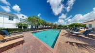Located at 6116 Yellow Sun Drive in Lantana, Florida, this outdoor amenity space showcases a well-maintained community swimming pool set against a backdrop of Florida's vibrant blue skies and lush tropical greenery. The rectangular pool features clear, turquoise water and is accessed via built-in entry steps, with safety life rings visibly stationed along the perimeter. The pool is framed by a generously sized paver deck constructed from warm-toned brick-style pavers, providing an attractive and durable surface for poolside activity. A row of lounge chairs and seating is positioned along one side of the deck, offering a comfortable space to relax and enjoy the South Florida sunshine. The surrounding landscape includes mature trees and manicured hedging that create a sense of privacy and a lushly shaded border around the area. A classic lamp post adds a charming architectural accent, while a neighboring multi-story building with a distinctive white facade is visible in the background, adding context to the community setting. The open-air environment benefits from abundant natural light, making this a bright and inviting outdoor retreat well-suited to the warm Florida climate.