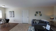 This single-floor space centers around a comfortable living room designed for both relaxation and everyday functionality. The room is finished with wall-to-wall carpeting complemented by a geometric-patterned area rug, creating a warm and inviting atmosphere underfoot. Neutral, light-toned walls provide a clean backdrop throughout, keeping the space feeling open and airy.

A prominent sliding glass door dominates one wall of the living room, fitted with vertical blinds that allow for adjustable light control. The door opens to an outdoor balcony, offering views of the surrounding exterior and allowing natural light to filter generously into the space during the day.

The living room accommodates a seating arrangement that includes a sofa with accent pillows, a standalone accent chair, and a low-profile coffee table positioned over the area rug. A media console with a flat-screen television is situated along one wall, providing a clear focal point for the room. Wall-mounted artwork and floor lamps contribute to the overall aesthetic. A paneled entry door with a standard hardware finish serves as the main access point into the unit.