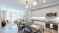Located at 7200 Baltimore Ave in College Park, Maryland, this single-floor residence offers a thoughtfully designed open-concept layout that seamlessly connects the kitchen, dining, and living areas. The contemporary kitchen is outfitted with sleek white cabinetry extending to the ceiling, a classic subway tile backsplash with under-cabinet lighting, dark countertops, and a full suite of stainless steel appliances. A central island with a dark countertop provides additional prep and dining space, illuminated by striking globe pendant lights. The open dining area flows naturally into the living room, which is anchored by large floor-to-ceiling windows and a glass sliding door that opens onto a private balcony, flooding the space with natural light and offering views of the surrounding neighborhood. Light hardwood flooring runs throughout, enhancing the bright, airy ambiance. The bedroom is generously proportioned with large windows, soft neutral tones, and ample space for a dedicated work area. The overall design aesthetic is clean and modern, with a warm, inviting atmosphere that balances contemporary finishes with comfortable living — ideal for those seeking a well-appointed residence in the heart of College Park.