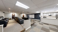Located at 2470 Southvale Crescent in Ottawa, Ontario, this dedicated entertainment space is thoughtfully designed to deliver an immersive cinematic experience. The main area is dominated by a tiered, stadium-style seating arrangement featuring rows of plush black leather power recliner chairs, each equipped with cup holders, offering every viewer an unobstructed sightline to the large wall-mounted projection screen at the front. A ceiling-mounted projector anchors the setup, while wall-mounted speakers and a subwoofer ensure quality surround sound. The room is finished with a neutral-toned drop ceiling fitted with recessed flat-panel light fixtures, and a bold navy blue accent wall frames the screen for a true theater aesthetic. A checkered carpet tile floor in varying shades of grey adds texture and helps with acoustics throughout the space. A low partition wall with a granite-topped ledge separates the viewing area from the entry, adding a refined architectural touch. Adjacent to the theater is a well-appointed kitchenette featuring white shaker cabinetry, granite countertops, a subway tile backsplash, stainless steel appliances including an electric range and microwave, a full-size refrigerator, and light wood-look tile flooring — ideal for entertaining guests before or during screenings.