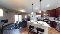 This single-floor space showcases a thoughtfully designed open-concept layout that seamlessly integrates the kitchen, dining, and living areas. The kitchen is appointed with rich dark wood cabinetry, stainless steel appliances including a French door refrigerator, microwave, and range, and a generous granite-topped island that doubles as a casual dining surface with two bar stools beneath it. Three sleek pendant lights hang above the island, adding both functional illumination and a modern aesthetic touch.

The kitchen flows naturally into the living area, which features warm hardwood flooring that extends throughout the space, creating visual continuity. The living area is anchored by a comfortable sofa and accented with a striped area rug, adding texture and definition to the space. A sliding glass door fills the living area with natural light and provides direct access to a private balcony, enhancing the sense of openness. Neutral wall tones complement both the cabinetry and flooring, while recessed lighting supplements the natural light throughout. Overall, the space offers a cohesive, functional, and inviting atmosphere.