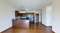 This single-floor unit is situated within a multi-story residential building distinguished by its contemporary exterior featuring yellow and neutral tones, individual balconies, and a well-appointed shared courtyard. The interior showcases an open-concept kitchen and living area with rich, dark hardwood-style flooring throughout. The kitchen is equipped with dark espresso cabinetry, a tile backsplash, and dark countertops that extend into a peninsula, providing additional prep space and a casual dining option. Black appliances, including a refrigerator, gas range with overhead microwave, and dishwasher, complete the functional kitchen. A pendant light fixture with multiple globe lights hangs above the peninsula, adding a decorative focal point. The open layout allows the kitchen to flow seamlessly into the living area, creating a cohesive and spacious feel. Natural light fills the space through well-positioned windows. The building's shared courtyard is a notable amenity, featuring a multi-tiered stone fountain as a centerpiece, mature landscaping with seasonal plantings, a pergola, and multiple seating areas with wrought-iron furniture and colorful accent chairs, offering residents an inviting outdoor gathering space.