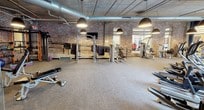 This fitness center combines industrial aesthetics with modern functionality across a single-floor layout divided into multiple workout zones. The space is defined by its exposed brick walls, concrete ceilings with visible ductwork, and bold navy blue accent walls, creating a dynamic, urban atmosphere throughout.

The cardio zone features large street-facing windows that flood the area with natural light and provide views of the surrounding neighborhood. Elliptical machines and treadmills are positioned to take advantage of these views. Oversized pendant lights hang from the ceiling, providing warm, focused illumination across the floor.

A dedicated strength training area offers a comprehensive selection of equipment, including cable machines, weight benches, resistance machines, and a power rack, all arranged on durable carpet flooring along the exposed brick wall. This section is further enhanced by additional pendant lighting.

A separate studio space, enclosed by glass partitions, provides a versatile area for stretching, yoga, and group fitness activities. This zone features dark stone-effect flooring, wood-paneled walls, a wall-mounted television, full-length mirrors, and decorative artwork, offering a more refined and focused environment distinct from the main floor.