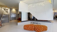 This contemporary single-floor space makes an immediate impression with its bold architectural design and refined aesthetic. The main area is defined by soaring ceilings that amplify the sense of openness and volume. A dramatic large-scale mirror panel spans a prominent wall, enhancing depth and light throughout the space. The ceiling is adorned with an artistic cluster pendant light installation, with delicate points of light arranged in an organic, branch-like pattern that serves as a stunning focal point overhead.

The interior features clean-lined white paneling across the walls and ceiling, creating a crisp, gallery-like atmosphere. A built-in bench seating area with a dark cushion runs along the base of the mirrored wall, complemented by a sleek glass coffee table. Vibrant orange ottoman seats with a geometric print pattern add a pop of color and a playful contrast to the otherwise neutral palette.

Adjacent to the main area, a secondary space is visible, featuring warm wood flooring, framed wall art, and classic red accent chairs, offering a more intimate seating environment. A stainless steel elevator is also present, suggesting a professional or upscale public-facing use. Light stone or natural material accents are visible in the background, adding textural richness to the overall design.