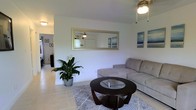 This single-floor residence offers a clean, comfortable living environment with a light and airy feel throughout. The living room serves as the heart of the home, featuring crisp white walls that enhance the sense of openness and brightness. Light-toned ceramic tile flooring runs throughout the space, providing a practical yet polished foundation that ties the rooms together seamlessly.

The living area is thoughtfully appointed, centered around a large sectional sofa in a neutral gray tone that pairs well with a round dark-wood coffee table accented with a glass insert. A decorative area rug anchors the seating arrangement, adding texture and warmth underfoot. A large framed wall mirror reflects natural light and contributes to the feeling of spaciousness, while coastal-themed artwork adds a subtle decorative touch to the walls.

A ceiling fan overhead ensures comfortable airflow throughout the main living space. Lush indoor greenery, including a prominently placed peace lily, introduces a fresh, natural element that enlivens the interior. A hallway visible from the living area connects the space to additional rooms, suggesting a functional and well-organized floor plan. Overall, the space presents a relaxed, inviting ambiance suited to comfortable everyday living.