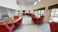 This elegant communal gathering space is designed to foster comfort and connection in a warm, classic setting. The room impresses immediately with its soaring double-height ceilings and crisp white walls, lending an open and airy atmosphere throughout. The patterned teal carpeting, accented by floral area rugs, adds a soft, traditional charm underfoot.

The main lounge area is thoughtfully arranged with multiple round wooden dining tables surrounded by rich red upholstered chairs, offering ample seating for socializing or group activities. A cozy seating nook near the fireplace features a stately black grand piano alongside a mounted flat-screen television above a classic white mantel with dark marble surround, flanked by a pair of pink wingback chairs — an inviting focal point for the room.

Large French doors with dark valances line one wall, flooding the interior with natural light and providing views of lush outdoor greenery. Adjacent to the main hall, a welcoming entry foyer features a round accent table, a decorative wrought-iron bench, and framed artwork, setting a refined tone upon arrival. Built-in cabinetry and classical architectural details complete the polished aesthetic of this thoughtfully appointed communal retreat.