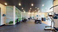 Located at 2020 Morton St in Charlotte, NC, this single-floor fitness facility is thoughtfully designed to accommodate a wide range of workout disciplines. The space features warm plywood wall paneling paired with dark ceilings and recessed lighting throughout, creating a contemporary yet functional aesthetic.

The main strength and conditioning area offers ample open floor space with durable rubber flooring, housing a cable machine, power rack, barbells, benches, and a dumbbell rack. Large windows along the perimeter flood the space with natural light and provide views of the surrounding greenery. Full-length mirrors are strategically placed to enhance visibility and spatial depth.

A dedicated functional training zone features unconventional equipment including a heavy tire, plyo boxes, and an air bike, catering to high-intensity and cross-training workouts. A separate cardio area includes rowing machines and additional cycling equipment positioned near glass partition walls.

The studio space stands out with its striking X-shaped architectural ceiling detail with integrated ambient lighting, polished floors, spin bikes, and yoga mats — ideal for group fitness classes. Rounding out the facility is a compact boxing room equipped with a heavy bag, pull-up bar, and direct outdoor access, all finished with the same warm plywood walls and dark rubber flooring.
