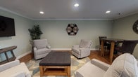 This thoughtfully designed single-floor space combines comfort and functionality in a cohesive, welcoming layout. The walls are finished in a soft sage green, complemented by crisp white crown molding and baseboards that run throughout, lending the space a clean, polished aesthetic. Warm hardwood flooring ties each area together seamlessly.

The main lounge area is generously sized and centers around a tufted dark gray ottoman coffee table, surrounded by multiple plush light gray swivel armchairs arranged atop a geometric-patterned area rug. A wall-mounted flat-screen television serves as a focal point on one end, flanked by a console table and a decorative potted plant. Recessed ceiling lighting provides even, ambient illumination throughout. A distinctive hexagonal wooden wall art piece adds a rustic, artistic accent to the space.

Adjacent to the lounge is a dining or gathering nook featuring a round high-top table with dark upholstered bar stools and a decorative wall clock, creating an informal setting for conversation or casual dining.

A dedicated workstation area is tucked along one wall, offering a granite-topped built-in desk with two computer monitors and bar-height seating for two. A window with horizontal blinds fills this area with natural light and garden views, making it a functional and pleasant space for focused work.