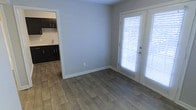 This single-floor space offers a clean, functional layout designed for comfortable everyday living. The main living area features warm wood-look flooring that extends throughout, complementing the soft, neutral gray walls and crisp white trim for a bright, modern aesthetic. Natural light flows generously into the space through a full-length exterior door fitted with white horizontal blinds, providing both privacy and access to the outdoors.

Adjacent to the living area, the kitchen is outfitted with dark espresso-toned cabinetry that creates a striking contrast against the lighter walls and flooring. The cabinetry provides ample storage, and the sink is conveniently positioned for practical use. The transition between the living and kitchen areas is seamless, lending an open and connected feel to the overall floor plan.

The space is characterized by its simplicity and clean lines, making it easy to personalize and furnish. White baseboards frame each room neatly, adding a polished finish to the interiors. The single-floor configuration ensures easy accessibility and effortless flow from one area to the next. Overall, this space presents a well-maintained, move-in-ready environment with a neutral palette suited to a variety of interior styles.