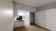 This single-floor studio is thoughtfully designed to maximize functionality within a compact footprint. The main living area features neutral-toned carpet flooring, soft gray walls, and a textured ceiling, creating a clean and understated aesthetic throughout. A standout feature of the living space is the built-in wall unit, which includes a fold-down Murphy bed, a built-in dresser with multiple drawers, and an adjacent closet with rod and shelf storage — offering smart, space-saving solutions without sacrificing practicality. A second open closet provides additional storage nearby. Natural light filters into the space through a window fitted with blinds, accompanied by a wall-mounted HVAC unit for year-round climate control. The kitchen is situated in an open alcove and features light wood-look vinyl flooring, dark granite-style countertops, white cabinetry, open shelving, a stainless steel range, and a black refrigerator — all accented by bright overhead lighting. A countertop peninsula connects the kitchen to the living area, enhancing the open flow. The bathroom is accessed through a doorway and includes a toilet, vanity with a dark countertop, and hardwood-style flooring, maintaining the cohesive neutral palette found throughout the unit.