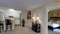 This single-floor residence offers a practical and comfortable layout designed for everyday living. The living area is finished with wall-to-wall carpet and neutral-toned walls, creating a warm and inviting atmosphere. Decorative wall art and a dark wood console table with accent lamps add a touch of character to the space, while a potted indoor plant near the kitchen entrance brings a natural element to the interior.

The kitchen is accessible directly from the living area through an open entryway, promoting a connected, open-plan feel. It features white cabinetry, light countertops, and tile flooring, providing a clean and functional workspace. Appliances are integrated within the cabinetry layout, and under-cabinet lighting enhances visibility and ambiance.

The textured ceiling runs throughout the space, contributing to the overall cohesive aesthetic. Natural and artificial lighting work together to keep the interiors bright and well-lit. The overall design is straightforward and functional, with a neutral color palette that offers flexibility for personalization. This space is well-suited for those seeking a comfortable, easy-to-maintain home with an efficient single-level floor plan.