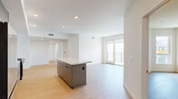 This thoughtfully designed single-floor residence showcases a clean, contemporary aesthetic throughout. The open-concept living and kitchen area forms the heart of the home, where light wood flooring and crisp white walls create a bright, airy atmosphere. Recessed lighting adds a polished finish overhead.

The kitchen is a standout feature, equipped with full-height matte gray cabinetry, stainless steel appliances including a gas range, dishwasher, and refrigerator, as well as a decorative geometric tile backsplash. A central island with a white countertop and built-in storage provides additional workspace and casual seating opportunities.

The living area flows seamlessly toward floor-to-ceiling glass doors that open to a private balcony, flooding the space with natural light and offering outdoor views of the surrounding architecture. Additional bedrooms also feature sliding glass door balcony access, reinforcing the indoor-outdoor connection throughout.

The primary bedroom suite includes a walk-in closet and a well-appointed en-suite bathroom featuring a dual-sink vanity with gray cabinetry, a large mirror, and a full soaking tub with tiled surround. Additional rooms are generously proportioned, maintaining the same consistent palette of light wood floors and neutral walls, offering versatile and functional living spaces.