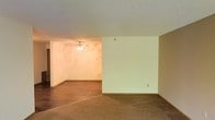 This single-floor space presents an open, functional layout that makes efficient use of its footprint. The main living area is finished with wall-to-wall carpet flooring, lending a warm and comfortable feel underfoot. Neutral, light-toned walls provide a clean backdrop throughout, allowing for versatile décor options.

A ceiling fan with an integrated light fixture is centrally positioned in the primary living area, offering both ambient lighting and air circulation year-round. The textured ceiling adds a subtle architectural detail that is consistent across the space.

The layout is defined by a clear division between areas — one section features smooth, off-white walls with standard electrical outlets, while an adjacent area showcases walls with a warm, decorative finish that adds visual interest and a touch of character. The transition between flooring materials — carpet in one zone and a darker hard surface in another — helps naturally delineate the different functional areas within the open plan.

Overall, the space offers a straightforward and adaptable interior, well-suited for a variety of uses, with its neutral palette and open configuration providing a solid foundation for personalization.